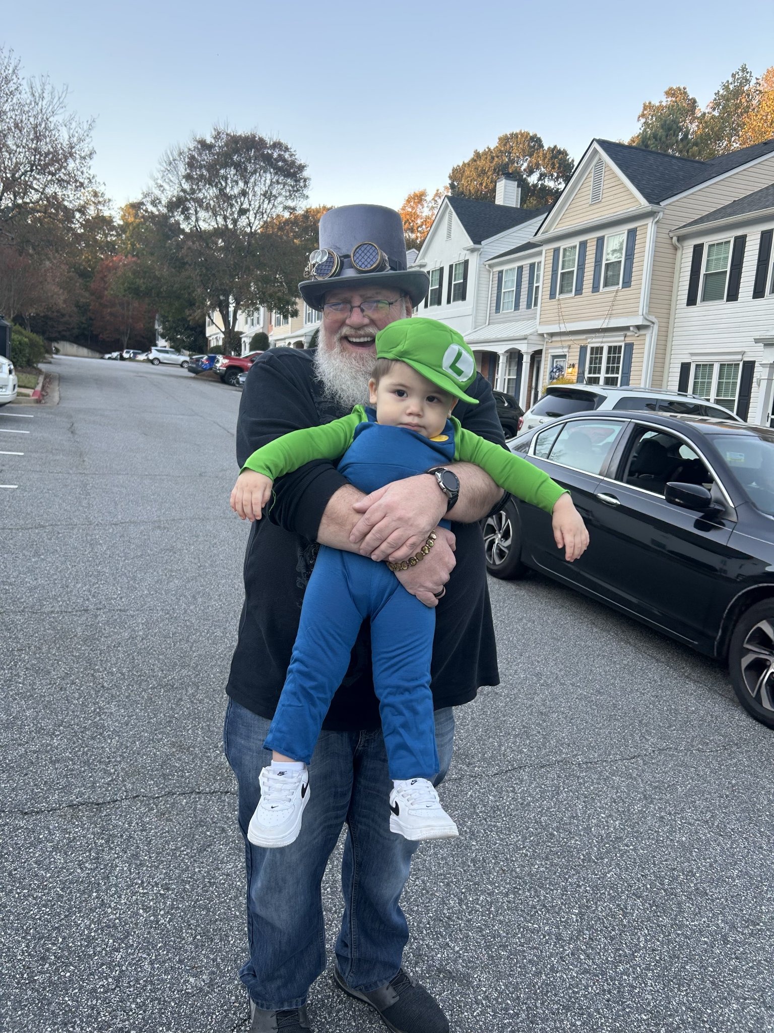 First Halloween trick-or-treating with my grandchildren — Jeffry in his steampunk top hat holding his Luigi-costumed grandson in the street, Fri · Oct 31, 2025